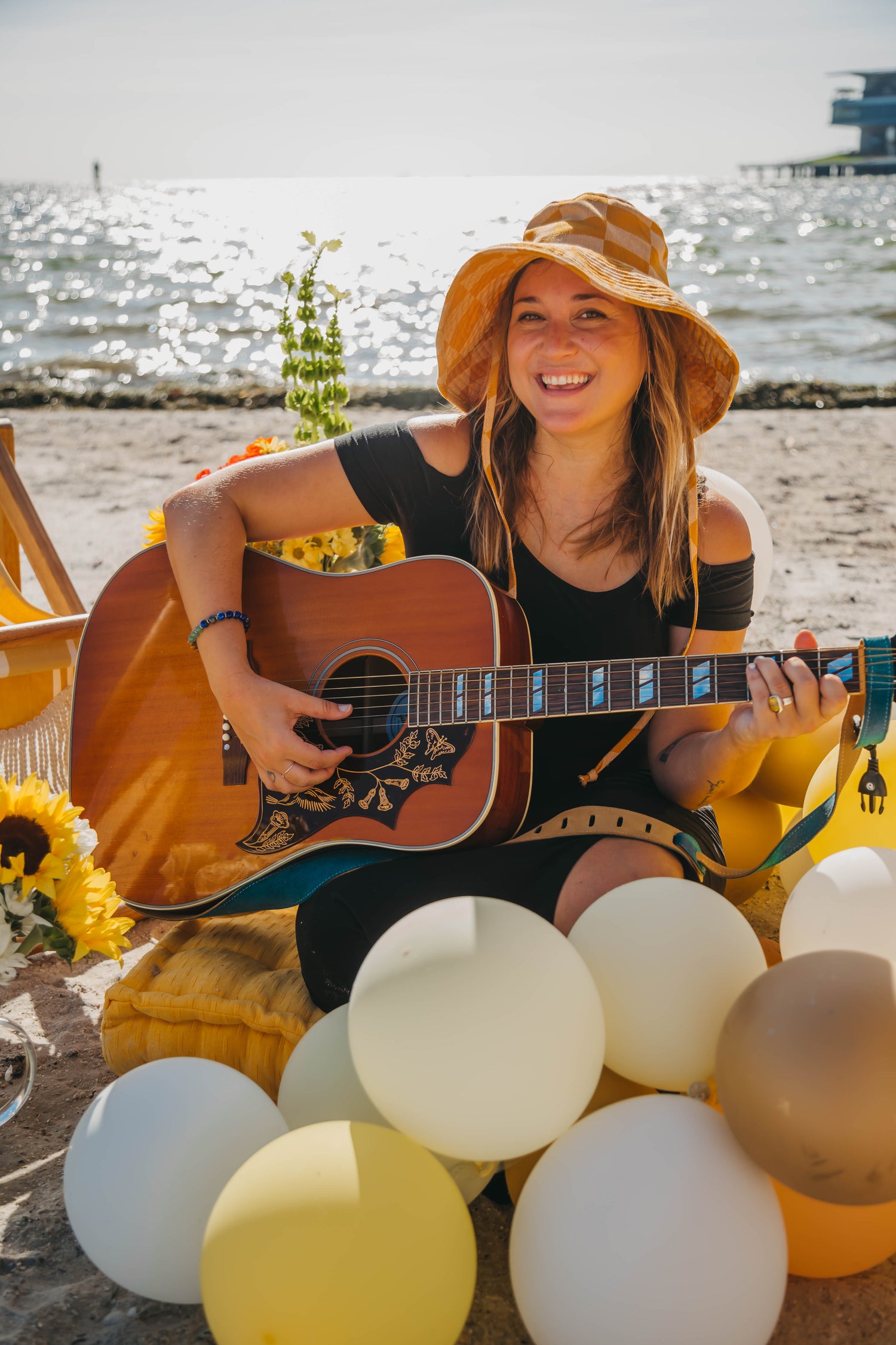 Photo of a girl wearing a sun hat sitting on a beach playing a guitar, and is surrounded by balloons and sunflowers.